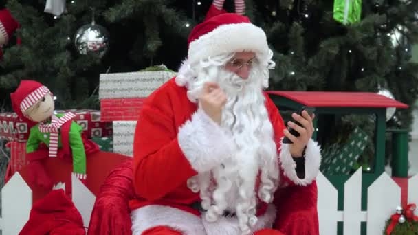 산타 클로스는 크리스마스 트리 근처 쇼핑몰에서 스마트폰으로 일하고 있다. — 비디오