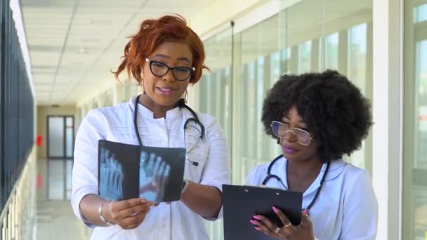 Duas médicas afro-americanas examinam o raio-X da perna, segurando-a nas mãos dentro de casa. Dois especialistas mantêm a imagem transparente da perna nos braços, e cuidadosamente a pesquisam — Vídeo de Stock