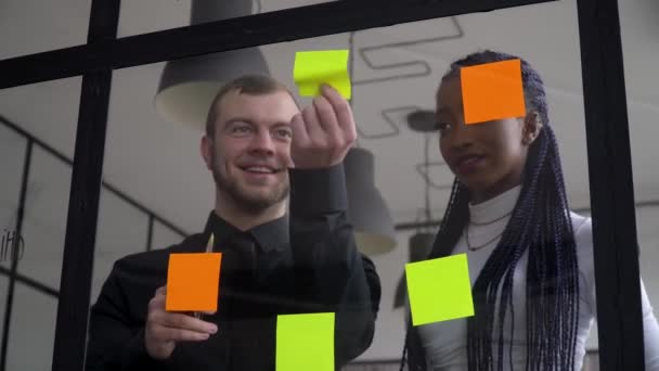 Företag företag multietniska människor med hjälp av klibbiga anteckningar, lösa strategi på glas whiteboard. Teamledare hane visar lösning för projektdeadline i kontorsmöte — Stockvideo
