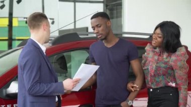 Genç yakışıklı araba satıcısı araba galerisindeki Afrikalı Amerikalı müşterilerine bir şey açıkladı. Otomobil işi, araba satışı, teknoloji ve insan konsepti