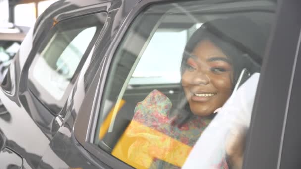 Glad afrikansk kvinna i en ny bil på ett stort affärsforum. Auto business, bilförsäljning, teknik och människor koncept. Bilen är svart — Stockvideo