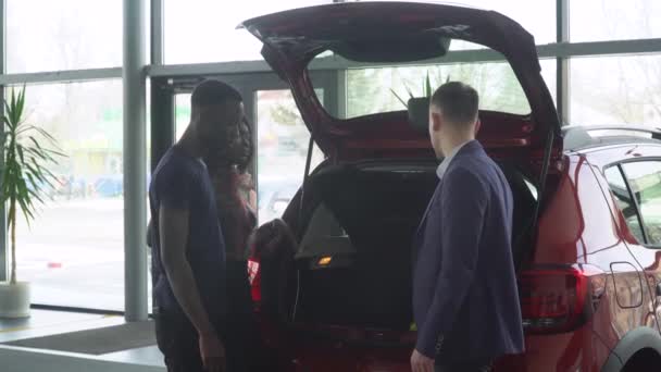 Vendedor de automóviles profesional está diciendo a los compradores interesados pareja afroamericana sobre coche de lujo con maletero abierto en el espectáculo del motor — Vídeo de stock