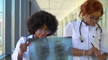 İki kadın Afro-Amerikalı doktor ciğerlerinin röntgenini çekiyorlar, içeride tutuyorlar. İki uzman göğsün şeffaf görüntüsünü kollarında tutuyor ve dikkatlice araştırıyor.
