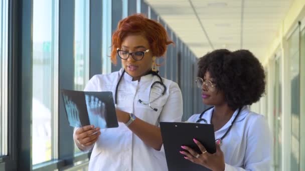 Duas médicas afro-americanas examinam o raio-X da perna, segurando-a nas mãos dentro de casa. Dois especialistas mantêm a imagem transparente da perna nos braços, e cuidadosamente a pesquisam — Vídeo de Stock