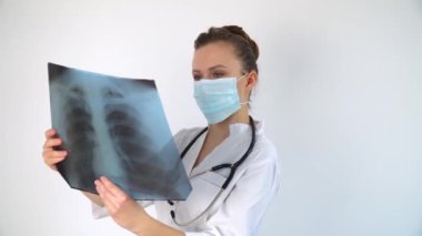 Koruyucu maskeli beyaz bir doktor, ciğerlerinin röntgenini inceleyip içeride tutuyor. Uzman, göğsün şeffaf görüntüsünü kollarında tutuyor ve Coronavirus için dikkatlice araştırıyor.