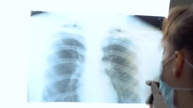 Koruyucu maskeli beyaz bir doktor, ciğerlerinin röntgenini inceleyip içeride tutuyor. Uzman, göğsün şeffaf görüntüsünü kollarında tutuyor ve Coronavirus için dikkatlice araştırıyor.