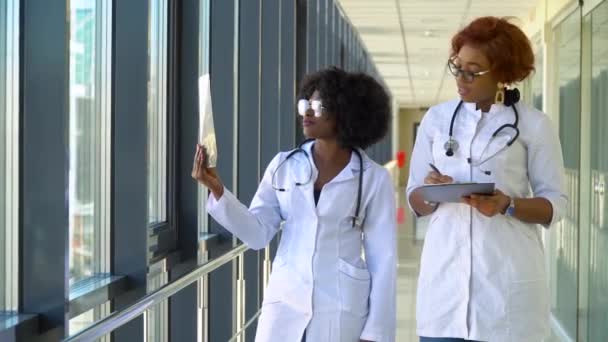 Két női afro-amerikai orvos vizsgálja a tüdő röntgenjét, és bent tartják a kezükben. Két specialista tartja átlátszó kép mellkas karok, és gondosan kutatja azt — Stock videók