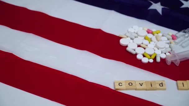 Слово коронавирус из букв на флаге США. Ковид - 19 вирусов. Таблетки и шприц на флаге США — стоковое видео