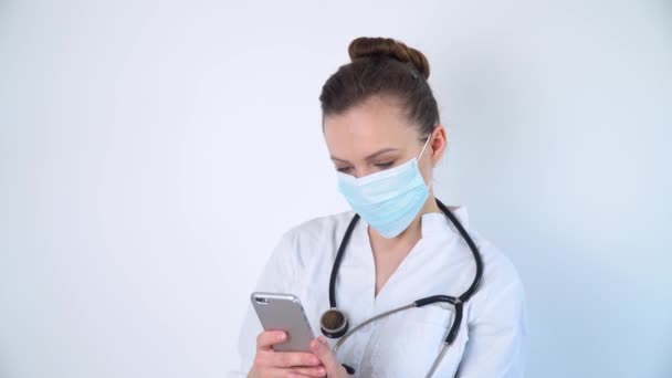 Jeune infirmière dans un masque de protection utilise son smartphone sur fond blanc dans un hôpital — Video