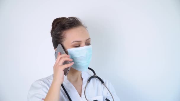 Koruyucu maskeli genç hemşire, hastanede beyaz bir arkaplanda telefonla konuşuyor. — Stok video
