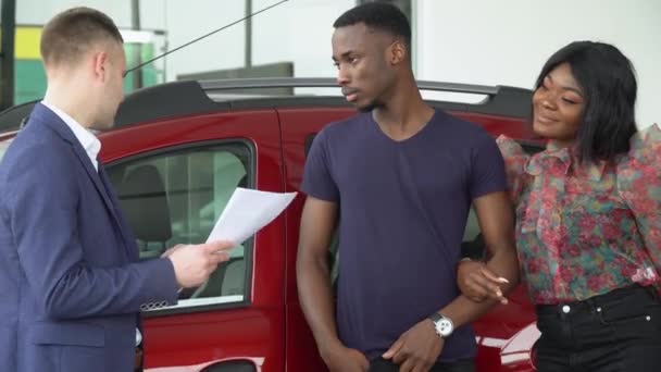 Jonge knappe autoverkoper legt iets uit aan zijn Afrikaanse Amerikaanse klanten bij de autodealer. Verklaring van de kenmerken — Stockvideo