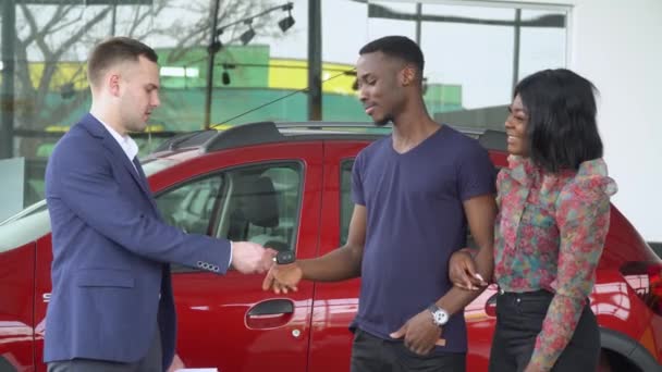 Молодий кавказький красивий продавець автомобілів дає своїм афроамериканським клієнтам ключі від нової машини — стокове відео