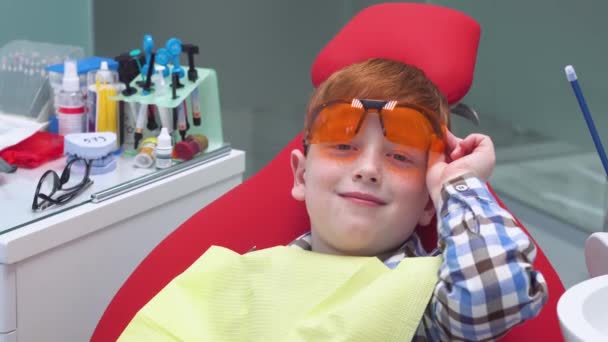Glücklicher Junge setzt Schutzbrille am Empfang beim Zahnarzt im Zahnarztstuhl auf. Kinderzahnheilkunde — Stockvideo