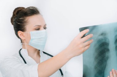 Koruyucu maskeli beyaz bir doktor, ciğerlerinin röntgenini inceleyip içeride tutuyor. Uzman, göğsün şeffaf görüntüsünü kollarında tutuyor ve Coronavirus için dikkatlice araştırıyor.