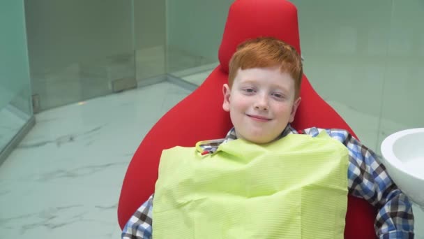 Gelukkige roodharige jongen bij de receptie bij tandarts in tandartsstoel dat is naar beneden. Pediatrische tandheelkunde — Stockvideo