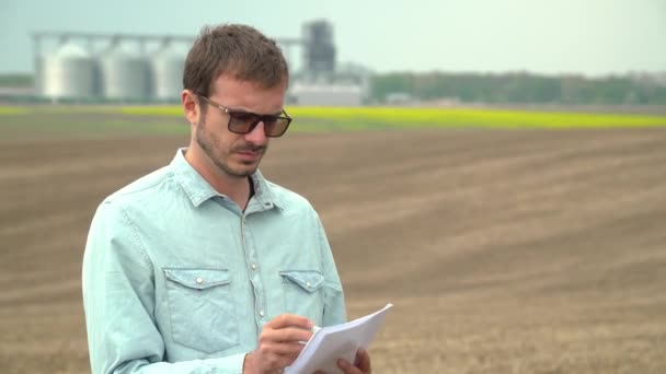 Landwirt kontrolliert das Feld vor Soja. Moderne Industrieanlage im Hintergrund — Stockvideo