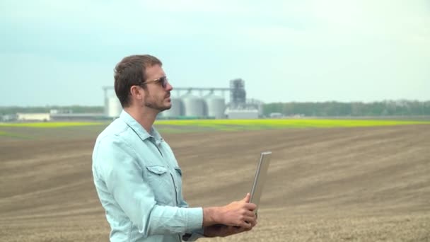 Laptoplu çiftçi soyadan önce tarlayı kontrol ediyor. Arka planda modern endüstriyel fabrika — Stok video