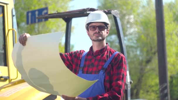 The worker in uniform looks at the drawings on the background of special road equipment — Stock Video