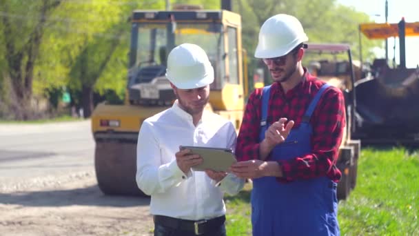 Gerente de los trabajos de reparación y trabajador en la carretera. El administrador de carreteras muestra el plan de construcción de carreteras de los trabajadores en una tableta. Concepto de reparación vial — Vídeos de Stock