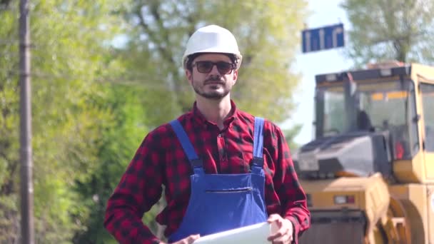 Arbeiter in Uniform und weißem Helm vor dem Hintergrund einer Straßenbaustelle blickt in die Kamera — Stockvideo