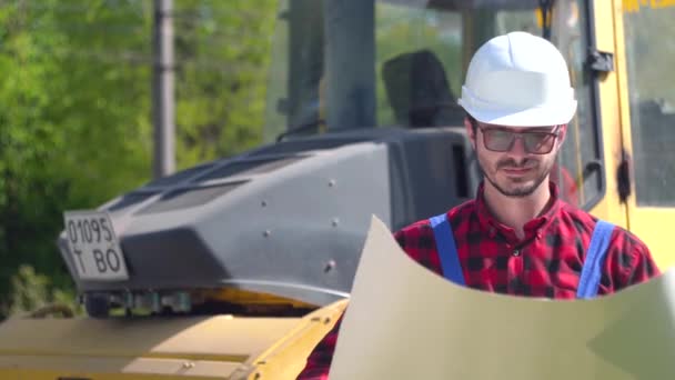 De werknemer in uniform kijkt naar de tekeningen op de achtergrond van de weg in aanbouw — Stockvideo