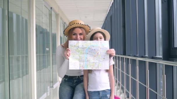 Beautiful woman with little girl holding a tourist map at the airport. Woman with daughter covers faces with tourist map — Stock Video