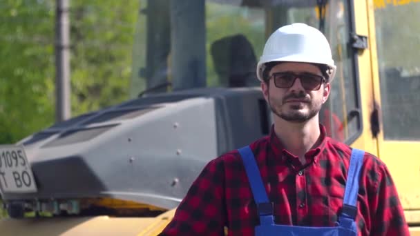 Porträtt av en byggare i hjälm och uniform mot bakgrund av en byggarbetsplats en solig sommardag. Vägreparationskoncept — Stockvideo