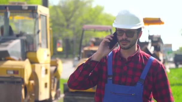 Bygga i hjälm och uniform mot bakgrund av en väg byggarbetsplats talar på telefonen — Stockvideo