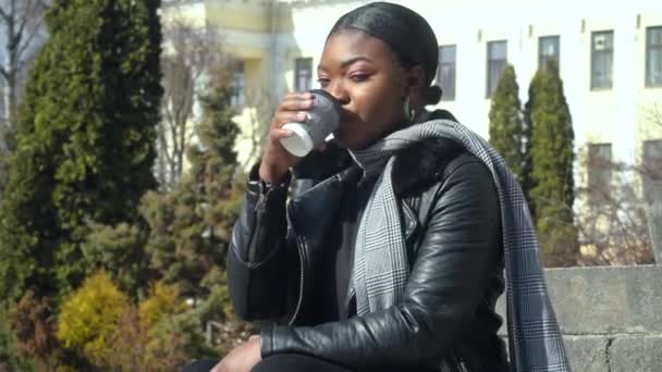 Glückliches junges afrikanisch-amerikanisches Mädchen, das Kaffee aus einer Papptasse trinkt, die auf der Treppe sitzt. Getränke- und Personenkonzept — Stockvideo