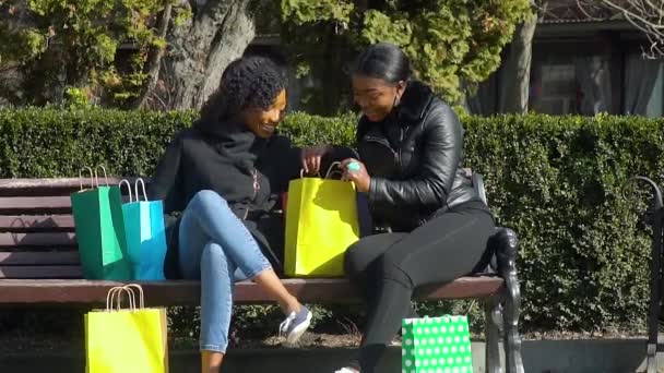 Две красивые африканские американские девушки получают удовольствие, сидя на скамейке со своими покупками после покупок — стоковое видео