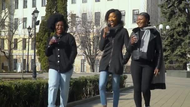 Three happy young african american girls drinking coffee from paper cup walking down the street — Stock Video