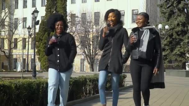 Tři šťastné mladé africké americké dívky pijící kávu z papírového kelímku kráčející po ulici