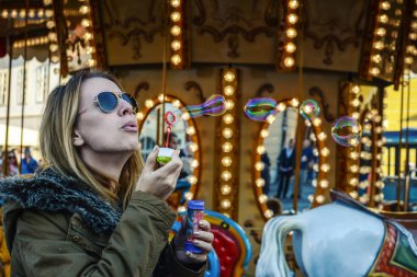 Atlıkarıncanın önünde baloncuklar çıkaran sarışın bir model. 