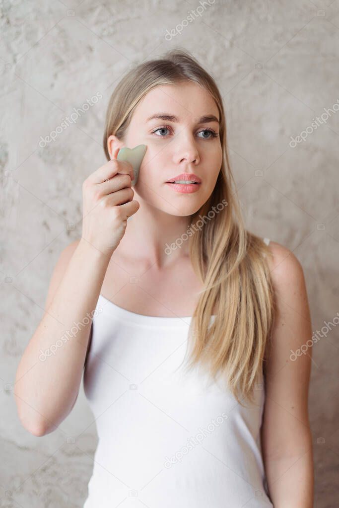 young blonde girl in a white t-shirt using facial gua sha jade board isolated at home on a beige background