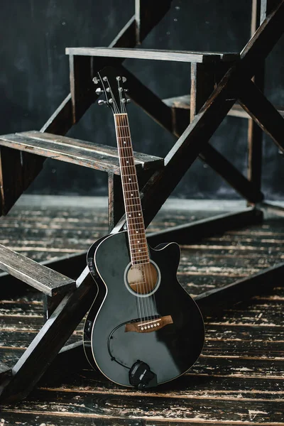 A black acoustic guitar stands in a room with a dark interior. Dark mood, loft, music, musical instrument.