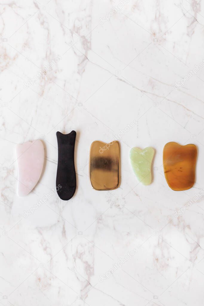 A variety of Guasha scrapers for facial massage and rejuvenation on a marble background. Rose quartz, black stone, jade.