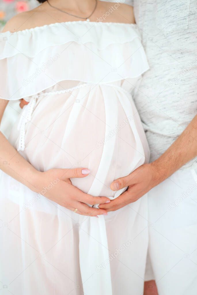 Pregnancy. A girl in a white peignoir with her husband is holding a stomach in their hands. Lightness and tenderness.