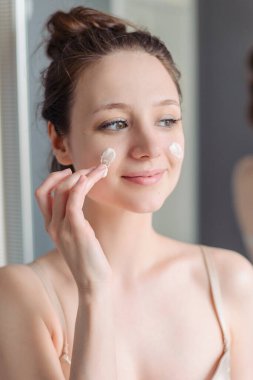 Young girl applies cream to face. Home light bathroom, lifestyle, natural skin care, love yourself. Cream on cheeks. A woman without makeup, natural. clipart