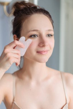 Kız evdeki hafif banyoda Gua sha masajı yapıyor. Gül kuvars masaj aleti, doğal taş. Kaldırma, doğal bakım.