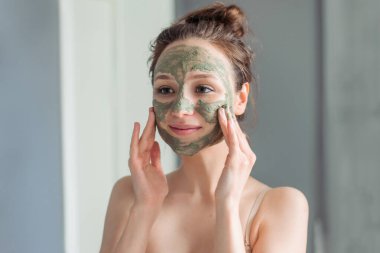 Girl applies a green mask of clay, minerals and salts to her face and looks in the mirror. Home light bathroom. Natural skin care. Vegetable vegan cosmetics. clipart