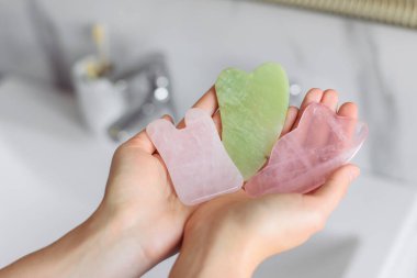 Guasha masseurs are in the hands of a girl on a bright white marble background in a home light bath. Green jade, rose quartz gua sha. Different forms of skin care scrapers. clipart