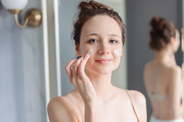 Young girl applies cream to face. Home light bathroom, lifestyle, natural skin care, love yourself. Cream on cheeks. A woman without makeup, natural.