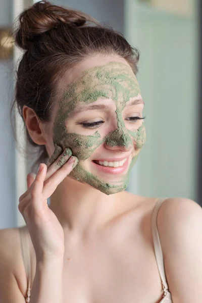Girl applies a green mask of clay, minerals and salts to her face. Home light bathroom. Natural skin care. Vegetable vegan cosmetics. Lovely young girl smiles.