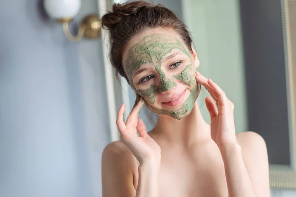 Girl applies a green mask of clay, minerals and salts to her face. Home light bathroom. Natural skin care. Vegetable vegan cosmetics. Lovely young girl smiles.