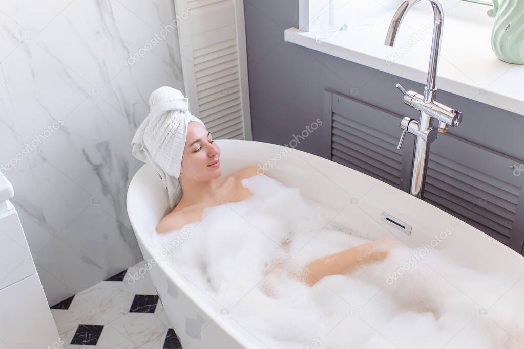 A young girl with a turban - a towel - on her head lies in a beautiful bright home bathroom and relapses. Daylight, marble tiles. The woman 's eyes are closed. Home spa and body care, lots of foam.