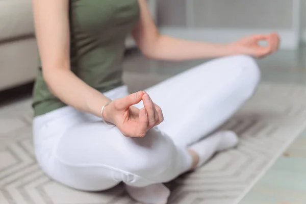 Woman does sports at home. White leggings and green top. Yoga, pilates, breathing, meditating, doing Ardha Padmasana exercise, Half Lotus pose with mudra gesture. Close up hands. Morning charging, workout, lifestyle.Well being, wellness concept.