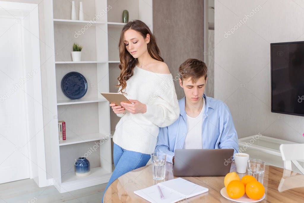 The young couple - male and female - work at home as freelance workers. The guy prints on the keyboard of the laptop computer, the girl holds the iPad. Home comfort, happy together.