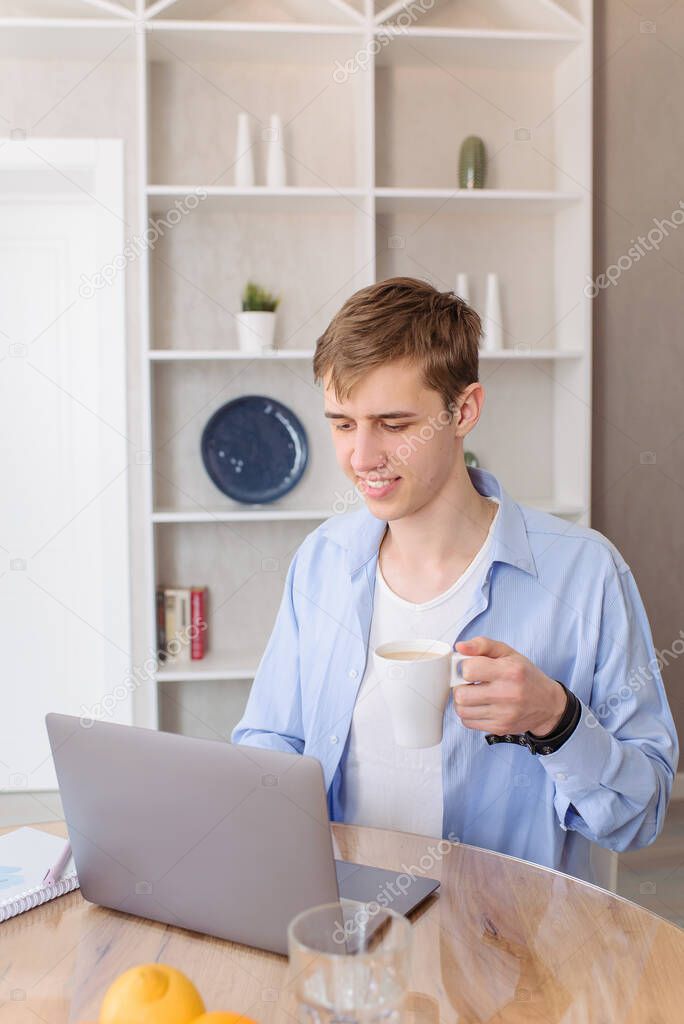 Young beautiful man works for a laptop computer from a home with a laptop as a freelancer. Weekend at house. Cup coffee in hand.
