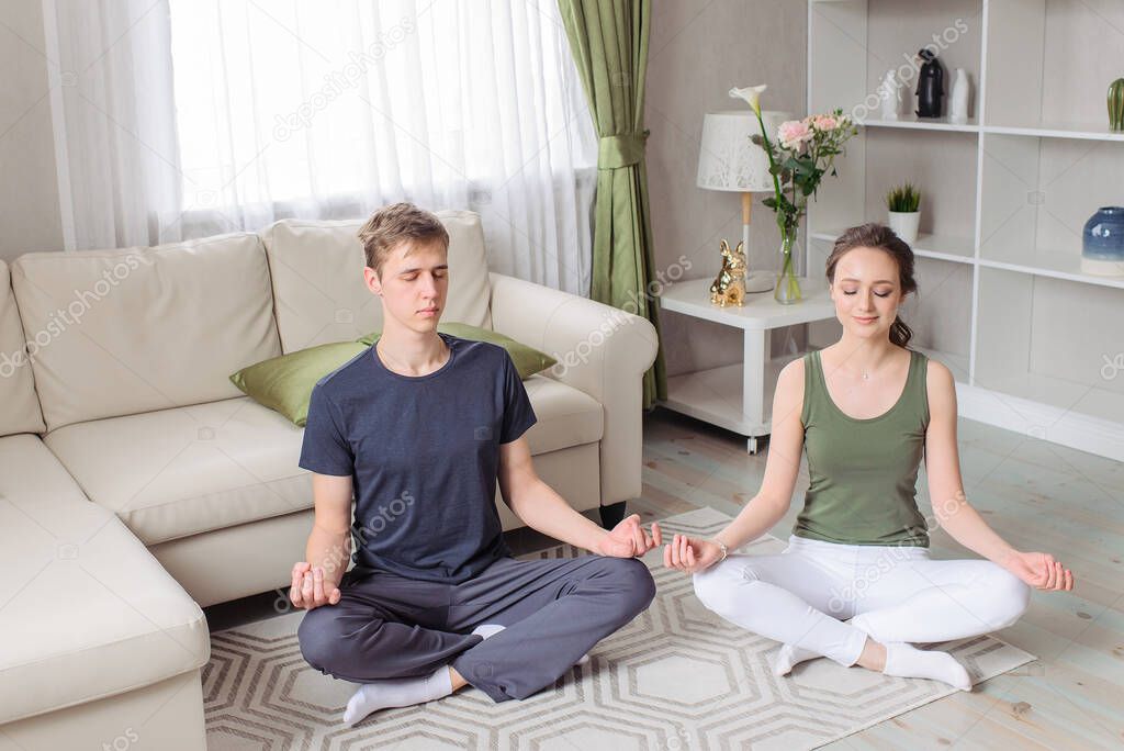 Young couple - man and woman - does sports at home. Sporting wear. Yoga, pilates, breathing, meditating, doing Ardha Padmasana exercise, Half Lotus pose with mudra gesture. Morning charging, workout, lifestyle.Well being, wellness concept.