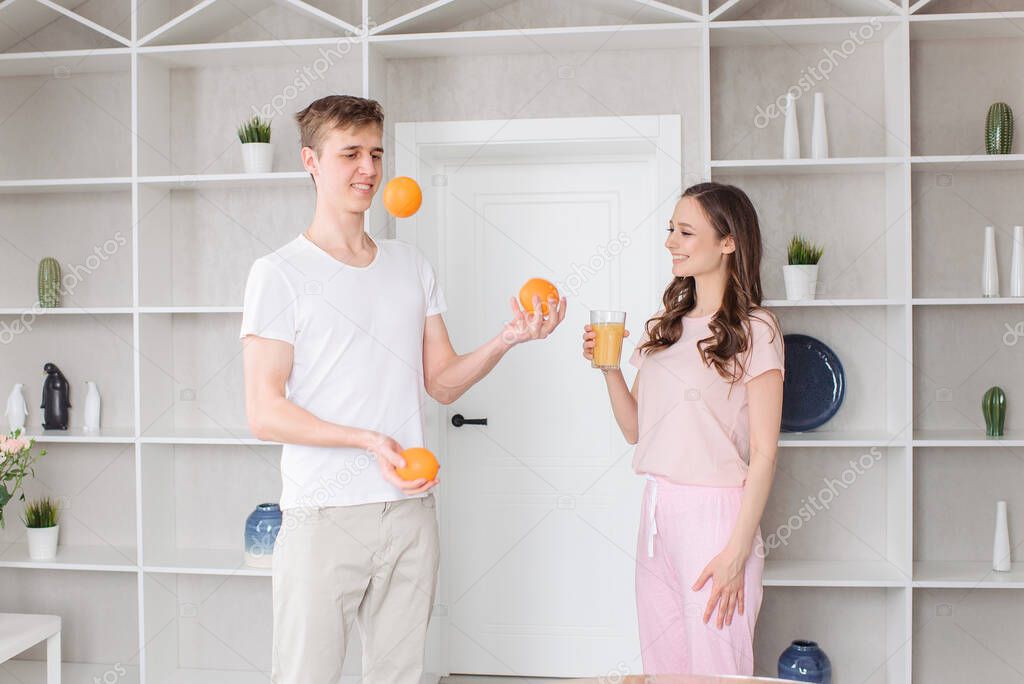 The guy juggles oranges, the girl laughs and drinks orange juice. A young couple in a cosy apartment.